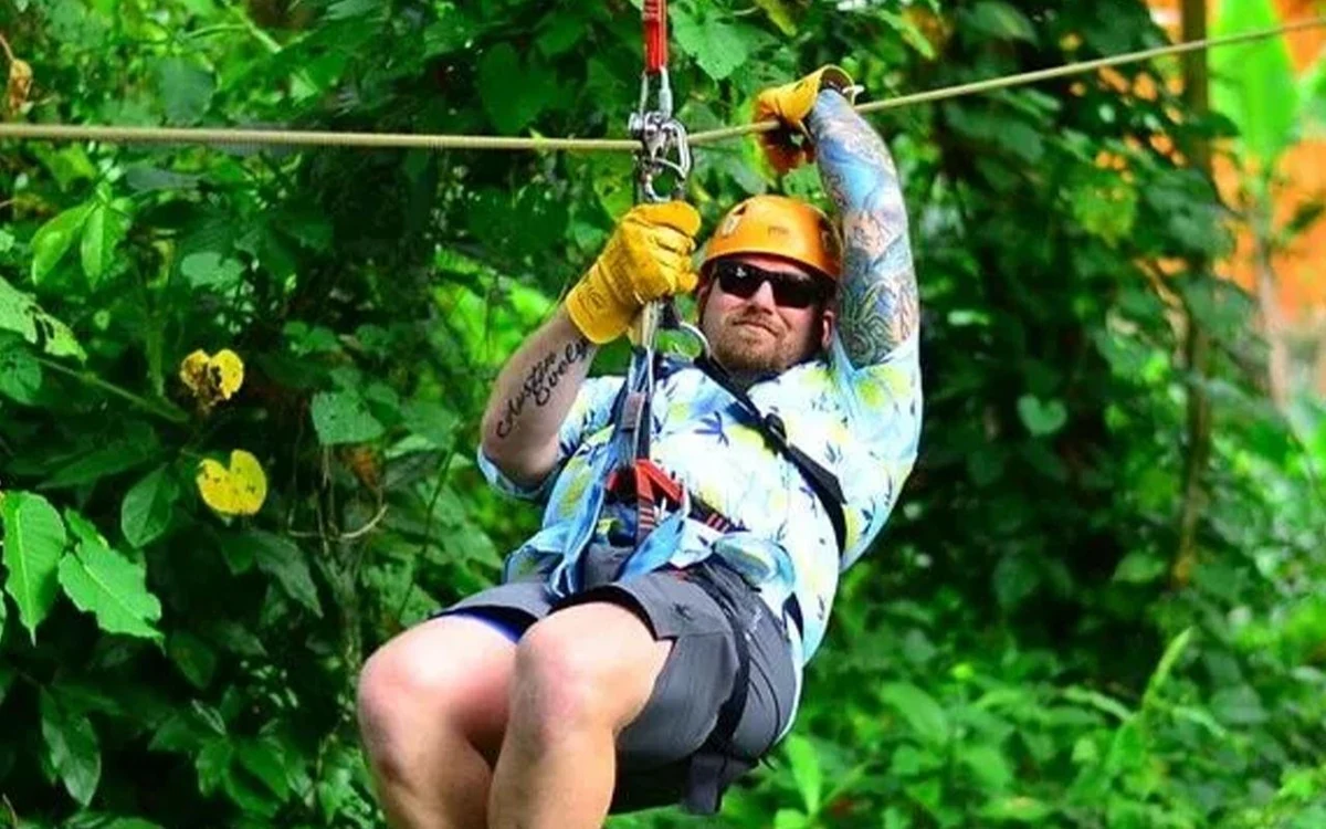 Ein Fahrer mit Helm und Gurtzeug gleitet an einer Seilbahn durch dichten grünen Dschungel