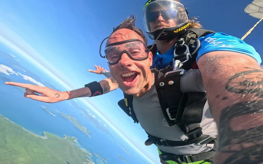 A tandem pair in freefall, the jumper grinning at the camera with the coast tilted below