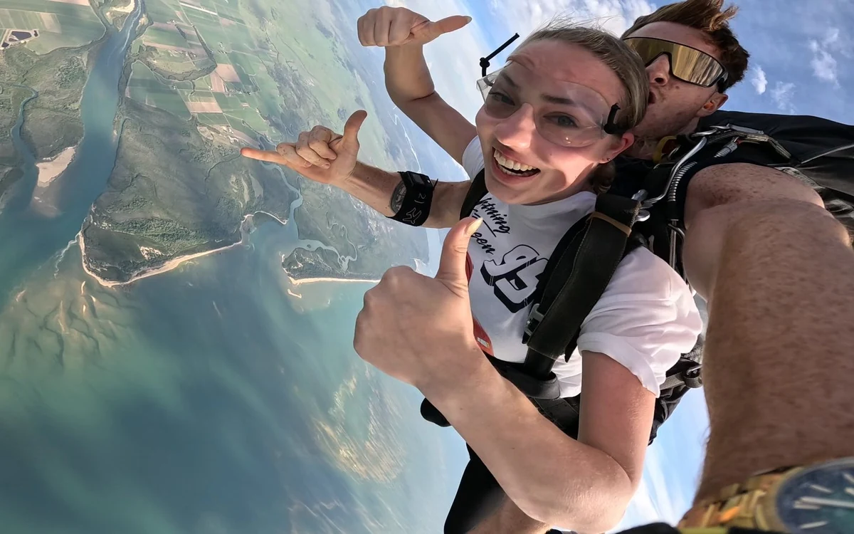 A tandem pair mid-air, with shallow turquoise water and a sandy headland behind them